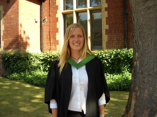 This is me, at 23, graduating from the University of Leeds. I have long blonde hair that is loose and a black graduation gown with a green collar. I'm standing outside a red brick university building.