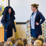 Energy Sparks team gives assembly. A child has been invited up to the front to demonstrate the effects of global warming, by piling jumpers on their head.