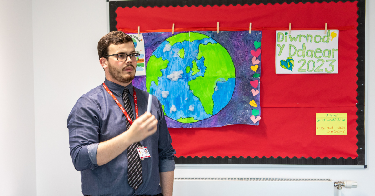 Secondary school teacher stands in front of a poster that says "Diwrnod y Ddaear 2023" ("Earth Day 2023"). The poster features a colourful painting of Earth.