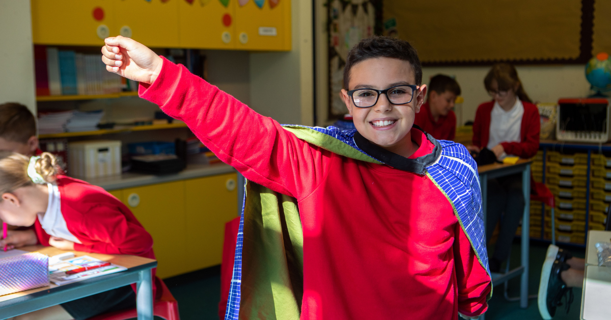 Primary pupil smiles to camera as he strikes a superhero pose. He is wearing a red jumper and a blue cape.