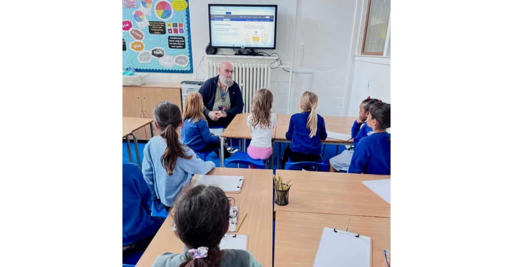 Image shows primary school students at Stillness Infant School speaking to their caretaker. A screen in the background shows the Energy Sparks scoreboard.