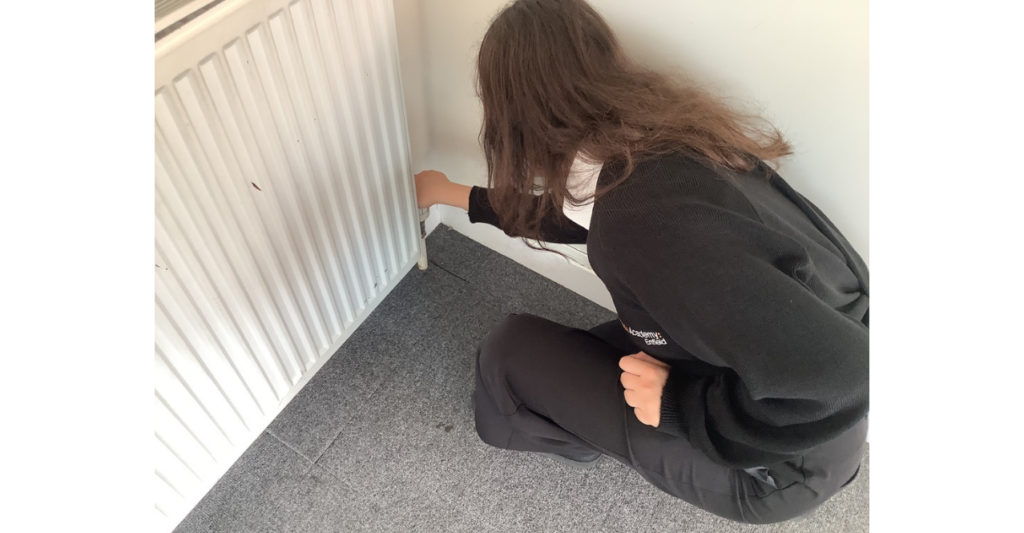 A secondary student crouches beside a radiator, adjusting the thermostat.